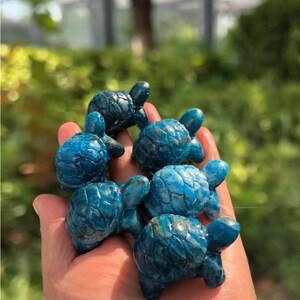 May include: A collection of small, blue turtle figurines. Each turtle has a detailed shell pattern and is a vibrant shade of blue. The figurines are held in a hand, with a blurred green background.