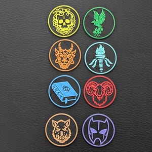 Può includere: Otto gettoni rotondi colorati con vari disegni, tra cui un teschio, una fenice, un drago, una torcia, un libro, un ariete, un cinghiale e una figura mascherata. Ogni gettone ha un bordo nero e uno sfondo di colore diverso.