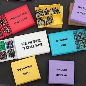 May include: A collection of rectangular plastic storage boxes with sliding lids, each labeled with names like Augmented Hoffman, Generic Tokens, Bygone Tiri, Infamous Zipp, and Brood Nekima. The boxes contain small, colorful game tokens in divided compartments.
