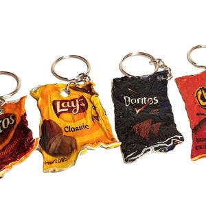 May include: Four novelty keyrings shaped like miniature snack food packets. The keyrings feature the logos of Fritos, Lay's Classic, Doritos, and Cheetos. Each keyring has a silver ring attached.