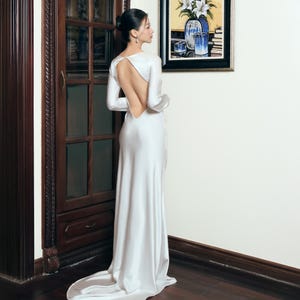 May include: A white satin gown with long sleeves and an open back. The dress has a floor-length skirt with a slight train. The model is standing near a dark wood cabinet and a painting with a floral arrangement.