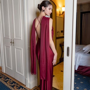 May include: A burgundy formal gown with a flowing cape detail. The dress has a low back and spaghetti straps. The model is standing in a doorway with a white door and a patterned rug.