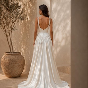 K&ouml;nnte beinhalten: Ein bodenlanges elfenbeinfarbenes Kleid mit tiefem R&uuml;ckenausschnitt, breiten Tr&auml;gern und einem figurbetonten Oberteil, das in einen volumin&ouml;sen, gefalteten Rock mit kleiner Schleppe &uuml;bergeht. Der R&uuml;cken des Oberteils ist mit einer Knopfleiste versehen.