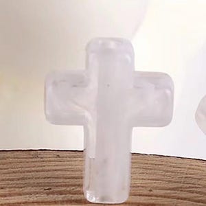 May include: A clear, translucent cross-shaped bead. The bead is made of a smooth, possibly crystal-like material. The cross is resting on a wooden surface, with a blurred background.