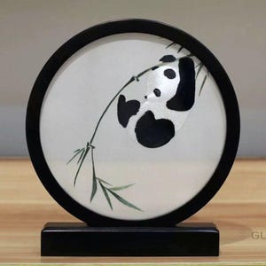 May include: A circular artwork in a black frame, depicting a panda bear on a bamboo branch. The panda is black and white, with green bamboo leaves. The artwork is mounted on a black stand.