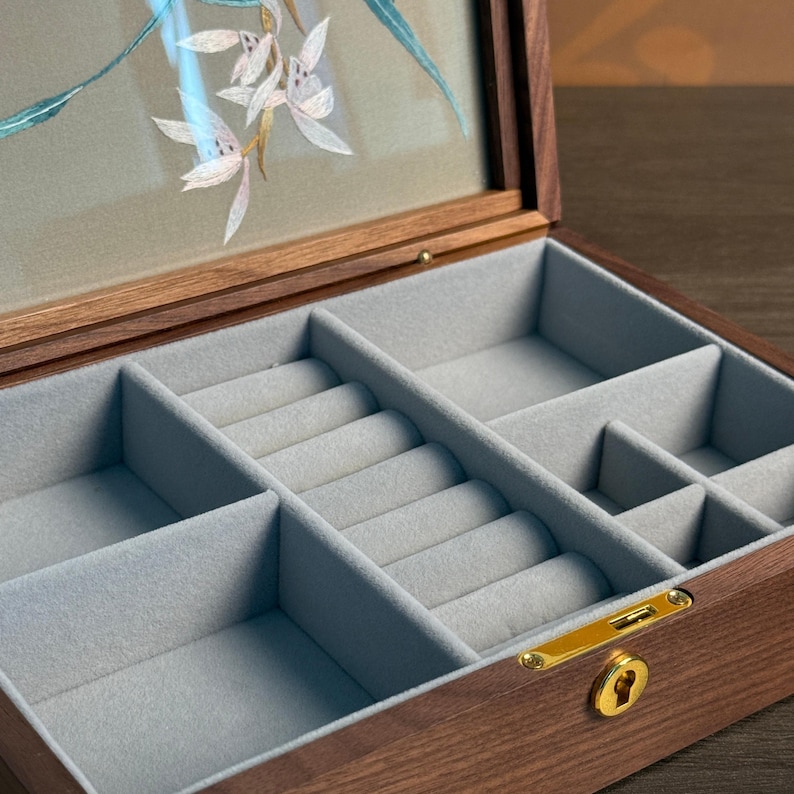 May include: A wooden jewelry box with a clear lid featuring a floral design. The interior is lined with gray felt and divided into compartments and ring slots. The box has a gold-colored clasp and keyhole.