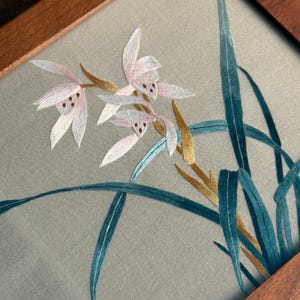 May include: Embroidered artwork featuring delicate pink and white flowers with maroon accents, gold stems, and teal leaves. The floral design is set against a gray background and framed in wood.