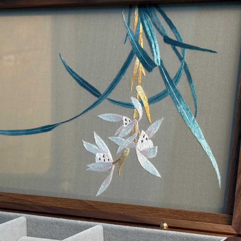 May include: A wooden jewelry box with a detailed embroidered floral design. The embroidery features light blue and white flowers with teal and gold accents. The box has a gray interior and a dark brown wooden frame.