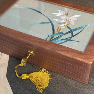May include: A wooden jewelry box with a glass top displaying an embroidered floral design in white, pink, and teal. The box has a gold-colored key and a yellow tassel. The wood has a rich, dark brown color.