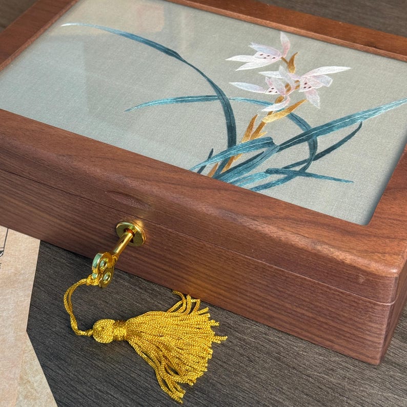 May include: A wooden jewelry box with a glass top displaying an embroidered floral design in white, pink, and teal. The box has a gold-colored key and a yellow tassel. The wood has a rich, dark brown color.