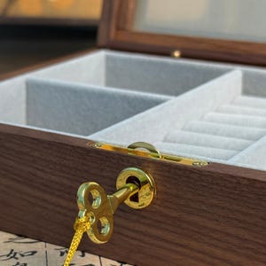 May include: A wooden jewelry box with a glass top and a gold-colored key. The interior is lined with a light gray fabric and has multiple compartments. The box is made of dark brown wood.