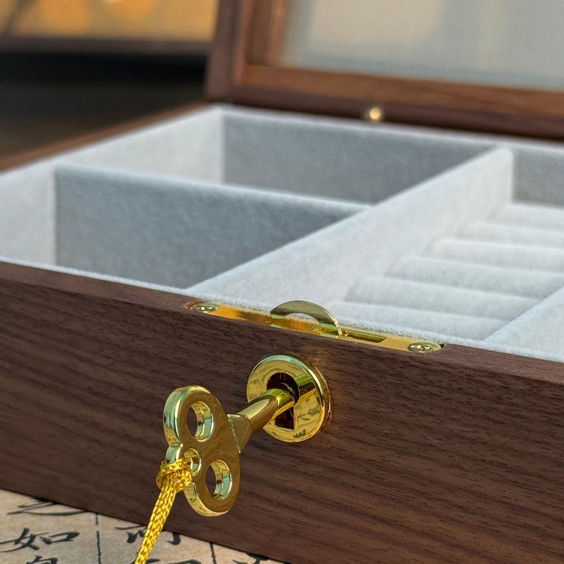 May include: A wooden jewelry box with a glass top and a gold-colored key. The interior is lined with a light gray fabric and has multiple compartments. The box is made of dark brown wood.