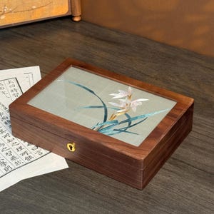 May include: A rectangular wooden box with a glass top, featuring an embroidered floral design in white, pink, and teal. The box has a gold-colored lock and is set on a dark wooden surface.