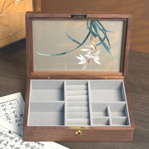 May include: Open wooden jewelry box with a light-colored interior and multiple compartments. The lid features an embroidered floral design with white flowers and teal leaves. The box is made of dark wood and has a gold-colored clasp.