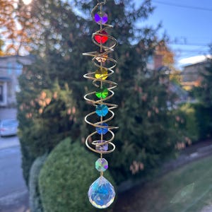May include: A hanging suncatcher with a gold-colored spiral design. It features a series of colorful, faceted crystal beads in purple, red, yellow, green, blue, and iridescent. A large, clear crystal ball hangs at the bottom.