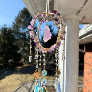 Puede incluir: Un atrapasoles colgante con un marco de metal dorado, adornado con cristales de amatista y colgantes de cristal transparentes e iridiscentes. El atrapasoles presenta un cristal de amatista central y detalles en forma de sol.