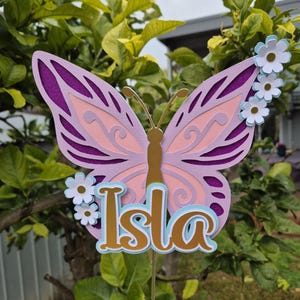 May include: A decorative butterfly made of layered paper in purple, pink, and gold, with the name "Isla" in gold and blue. The butterfly features intricate cutouts and is decorated with small white and blue flowers. It is mounted on a thin metal stake.