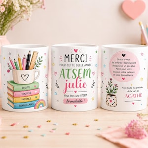 Può includere: Tre tazze in ceramica bianca con disegni colorati. Una tazza presenta matite, un righello e un cuore su una pila di libri. Un'altra tazza dice "Merci" e "ATSEM Julie". La terza tazza ha un messaggio e una pianta in vaso.