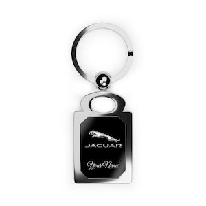 Könnte beinhalten: Ein silberfarbener, rechteckiger Schlüsselanhänger mit einem schwarzen Mittelteil, der das Jaguar-Logo und den Text "JAGUAR" und "Your Name" zeigt. Der Schlüsselanhänger hat einen kreisförmigen Schlüsselring und ein kleines dekoratives Element.
