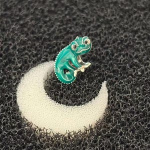 May include: A teal chameleon-shaped brooch with silver-colored trim and accents. The brooch is set on a white crescent moon shape, against a dark textured background. The chameleon has large, round eyes.