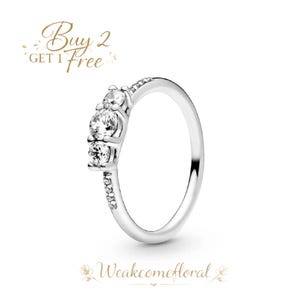Könnte beinhalten: Ein silberner Ring mit drei klaren, runden Edelsteinen in einer Reihe. Das Band ist ebenfalls mit kleineren, klaren Steinen besetzt. Der Ring ist vor einem weißen Hintergrund platziert. Der Text "Buy 2 Get 1 Free" befindet sich oben links.