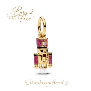 Puede incluir: Un charm dorado con forma de cascanueces soldado. El cascanueces tiene un sombrero y brazos rojos brillantes, con una estrella y una gema transparente. El charm tiene botas blancas y detalles en negro. El texto "Buy 2 Get 1 Free" está en la esquina superior izquierda.