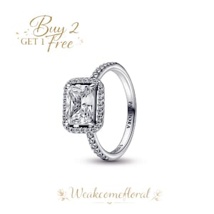 Peut inclure: Une bague argentée ornée d'une pierre précieuse rectangulaire et transparente, entourée de petites pierres transparentes. La monture de la bague est également ornée de petites pierres transparentes. Le mot "PANDORA" est visible sur la monture. Texte : "Buy 2 Get 1 Free".