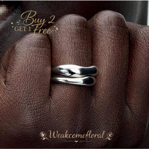 May include: A silver-colored ring with a unique, overlapping design. The ring is shown on a dark brown hand. The text "Buy 2 Get 1 Free" is in the upper left corner, and the text "Weakcomefloral" is at the bottom.