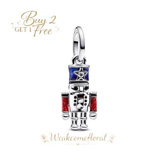 May include: A silver charm in the shape of a toy soldier nutcracker. The nutcracker has a blue and red hat and arms, with a star and a clear gem. The charm has a loop at the top for a necklace or bracelet. The text "Buy 2 Get 1 Free" is in the upper left corner.