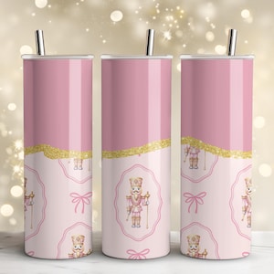 Könnte beinhalten: Drei rosa isolierte Becher mit silbernen Strohhalmen. Jeder Becher hat ein goldglitzerndes Band und ein hellrosa ovales Design mit einer Nussknacker-Illustration. Unter dem Oval befindet sich eine rosa Schleife. Der obere Teil der Becher ist einfarbig rosa.