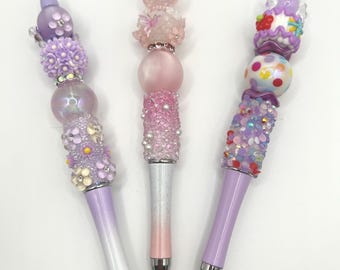May include: Three decorative pens with embellished designs. One pen features purple and white floral accents, another has pink beads and a rose, and the third has a cake-like topper with colorful gems. Each pen has a silver tip.