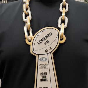 May include: A large, gold and silver chain necklace with a football-shaped pendant. The pendant reads "Lorenzo #14 4-0" and "2025 Toy Bowl 8th Grade Champions" with "LYF Lawrence Youth Football" text.