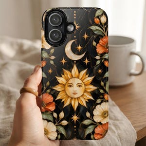 May include: A black smartphone case featuring a central golden sun face with rays, a crescent moon above, and scattered four-pointed stars. The design is surrounded by orange, white, and cream-colored flowers with green leaves on a dark background.