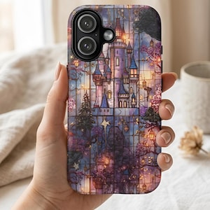 Pode incluir: Uma capa de telefone com um design de vitral de um castelo de conto de fadas com torres, árvores e lanternas. A capa tem um fundo escuro com tons roxos, azuis e dourados. A capa do telefone tem uma câmera dupla.