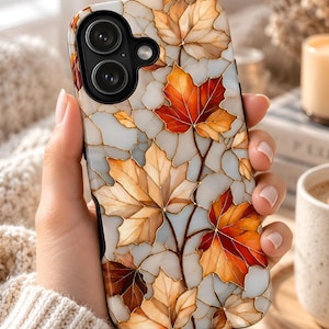 May include: A smartphone case featuring a stained-glass style design of autumn maple leaves in shades of orange, red, and brown against a white background with gold-colored outlines, surrounding a black camera module with two circular lenses.
