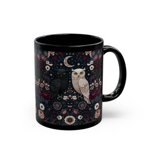 Puede incluir: Una taza de cerámica negra con una ilustración detallada de un búho oscuro y uno claro posados entre un denso patrón de flores coloridas, hojas y pequeñas estrellas, centrados bajo una luna creciente sobre un fondo azul oscuro.