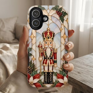 May include: A smartphone case featuring a stained glass style design of a nutcracker soldier in a red and gold uniform. The nutcracker holds a staff and is surrounded by holly leaves, red berries, and white poinsettia flowers on a light background.