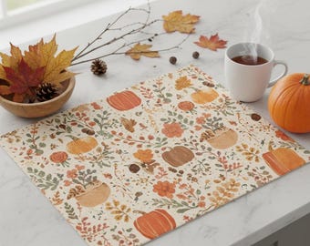 May include: A cream-coloured rectangular placemat with a pattern of orange pumpkins, flowers, and leaves. A white mug with a hot drink, a small pumpkin, and a wooden bowl with autumn leaves and pine cones are also visible.