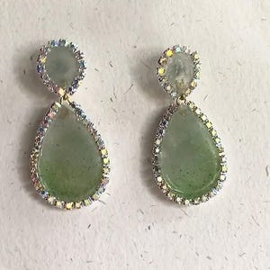 May include: A pair of green teardrop-shaped earrings with a clear crystal halo around each stone.