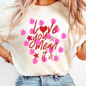 May include: Cream-colored t-shirt with the phrase "Love You Mean It" in red script, a red heart, and pink polka dots and stars. The shirt is a casual style.