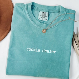 Pu&ograve; includere: Una maglietta Comfort Colors turchese con la scritta "cookie dealer" stampata in bianco. Una collana d'oro con un ciondolo poggia sulla maglietta. La maglietta &egrave; piegata e un cappello marrone &egrave; visibile sullo sfondo.
