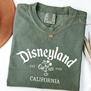 Może przedstawiać: Oliwkowo-zielona koszulka z napisem "Disneyland" w białym skrypcie, wraz z Myszka Miki i tekstem "Est. 1955 California". Złoty naszyjnik z wisiorkiem spoczywa na koszulce. Widoczny jest r&oacute;wnież brązowy kapelusz i suszone liście.