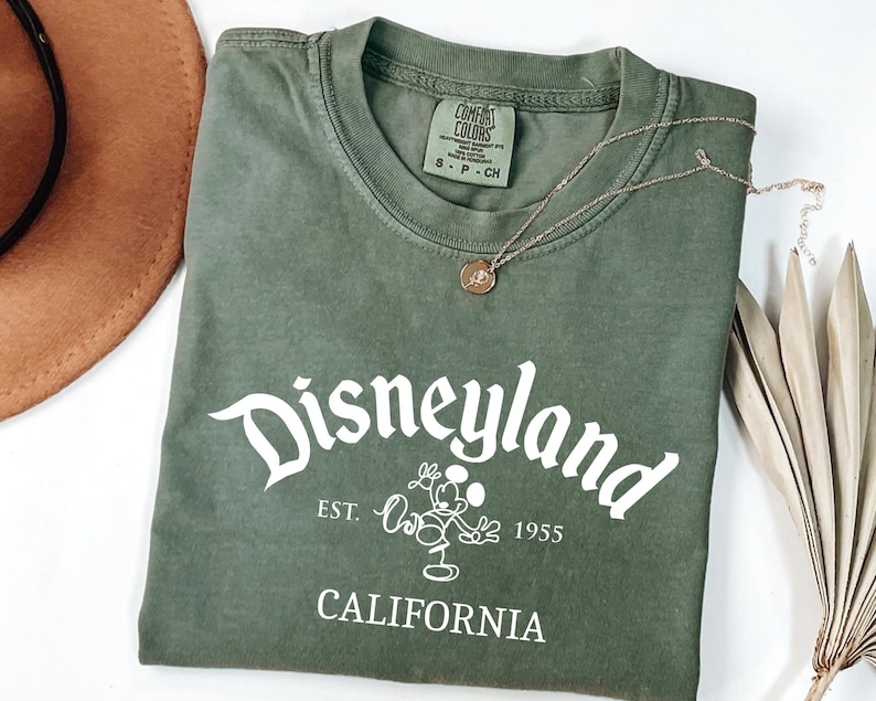 Może przedstawiać: Oliwkowo-zielona koszulka z napisem "Disneyland" w białym skrypcie, wraz z Myszka Miki i tekstem "Est. 1955 California". Złoty naszyjnik z wisiorkiem spoczywa na koszulce. Widoczny jest r&oacute;wnież brązowy kapelusz i suszone liście.