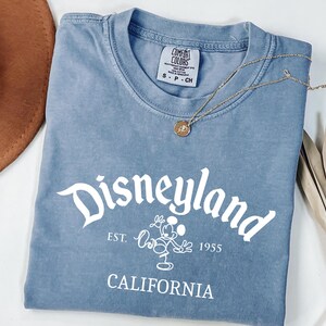 Może przedstawiać: Niebieska koszulka z białym napisem "Disneyland", a także "Est. 1955" i "California". Złoty naszyjnik z wisiorkiem spoczywa na koszulce. Brązowy kapelusz i suszona roślina znajdują się w tle.