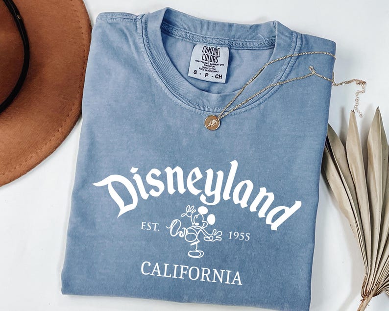 Może przedstawiać: Niebieska koszulka z białym napisem "Disneyland", a także "Est. 1955" i "California". Złoty naszyjnik z wisiorkiem spoczywa na koszulce. Brązowy kapelusz i suszona roślina znajdują się w tle.
