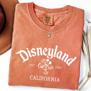 Może przedstawiać: Koralowa koszulka z logo Disneyland w kolorze białym, z Myszka Miki. Koszulka zawiera napis "Est. 1955" i "California". Widoczny jest r&oacute;wnież złoty naszyjnik i brązowy kapelusz.
