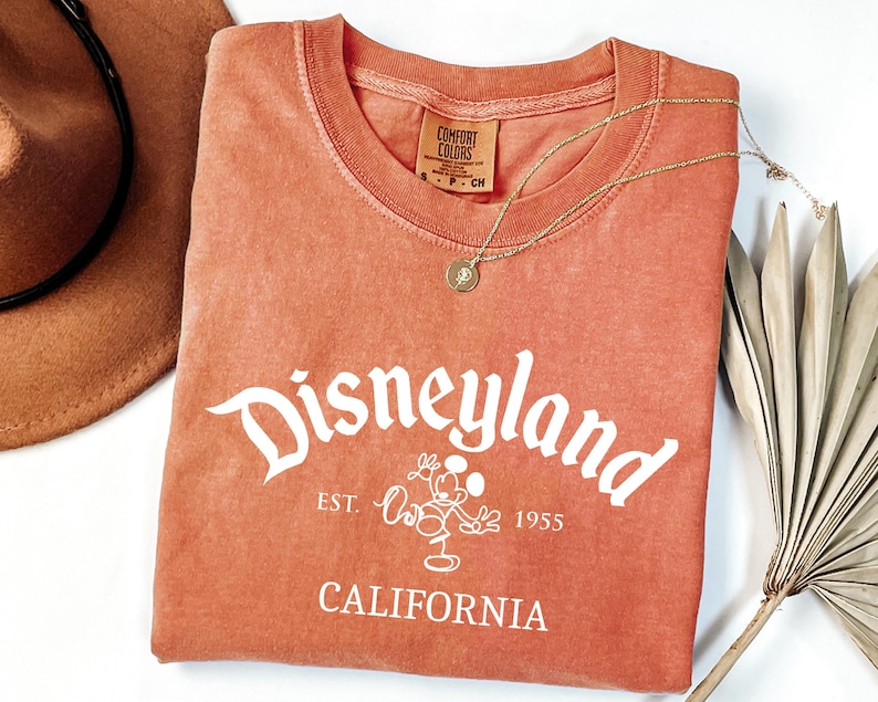Może przedstawiać: Koralowa koszulka z logo Disneyland w kolorze białym, z Myszka Miki. Koszulka zawiera napis "Est. 1955" i "California". Widoczny jest r&oacute;wnież złoty naszyjnik i brązowy kapelusz.
