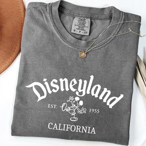 Może przedstawiać: Szara koszulka z napisem "Disneyland" w białym skrypcie, wraz z tekstem "EST. 1955 CALIFORNIA" i grafiką Myszki Miki. Widoczny jest r&oacute;wnież złoty naszyjnik i brązowy kapelusz.
