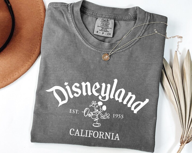 Może przedstawiać: Szara koszulka z napisem "Disneyland" w białym skrypcie, wraz z tekstem "EST. 1955 CALIFORNIA" i grafiką Myszki Miki. Widoczny jest r&oacute;wnież złoty naszyjnik i brązowy kapelusz.