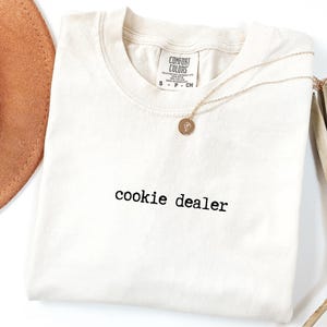 Pu&ograve; includere: T-shirt color crema con la scritta "cookie dealer" stampata in nero. Una collana d'oro con un ciondolo circolare &egrave; appoggiata sulla maglietta. Un cappello marrone e steli di piante secche sullo sfondo.